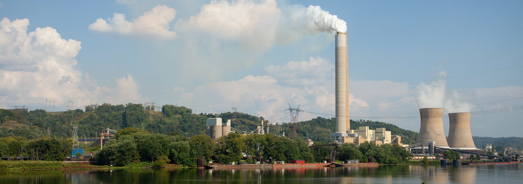 Power plant with a tall smokestack and cooling towers set against a hilly, green landscape